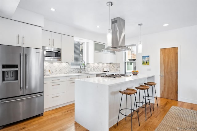 kitchen with backsplash, stainless steel appliances, island range hood, white cabinets, and decorative light fixtures