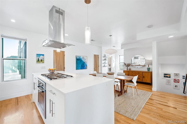 kitchen with light wood-type flooring, appliances with stainless steel finishes, pendant lighting, island exhaust hood, and white cabinets