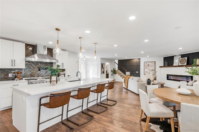 kitchen with white cabinets, wall chimney range hood, hanging light fixtures, and an island with sink