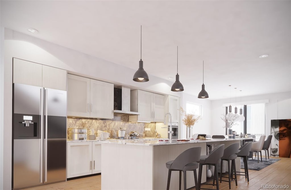 kitchen with stainless steel fridge, wall chimney exhaust hood, an island with sink, tasteful backsplash, and light wood finished floors