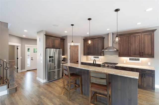 kitchen featuring light stone counters, a center island with sink, premium appliances, pendant lighting, and wall chimney range hood