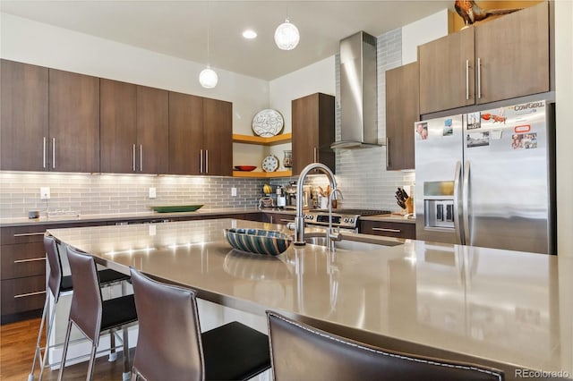 kitchen with pendant lighting, stainless steel fridge, a breakfast bar area, a kitchen island with sink, and wall chimney exhaust hood