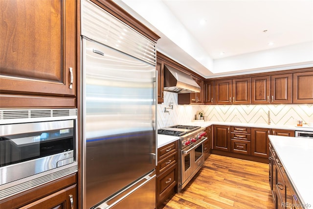 kitchen with built in appliances, custom exhaust hood, decorative backsplash, and light hardwood / wood-style floors