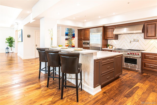 kitchen with built in appliances, tasteful backsplash, a kitchen island, hardwood / wood-style flooring, and wall chimney exhaust hood