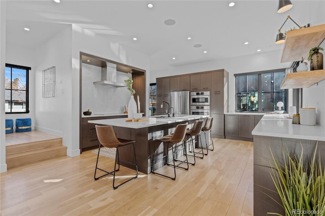 kitchen with appliances with stainless steel finishes, dark brown cabinets, a kitchen breakfast bar, an island with sink, and wall chimney exhaust hood