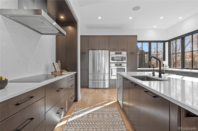 kitchen featuring wall chimney range hood, sink, appliances with stainless steel finishes, dark brown cabinetry, and light hardwood / wood-style floors