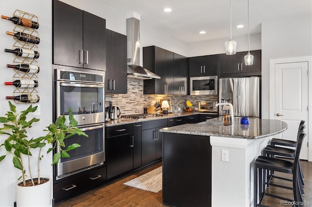 kitchen featuring wall chimney range hood, a breakfast bar area, dark cabinetry, and stainless steel appliances
