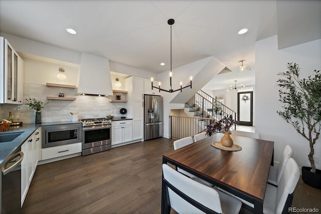 kitchen featuring dark countertops, appliances with stainless steel finishes, exhaust hood, a notable chandelier, and open shelves