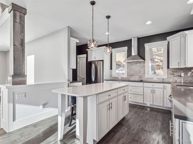 kitchen featuring wall chimney exhaust hood, dark wood-type flooring, pendant lighting, a kitchen island, and stainless steel refrigerator