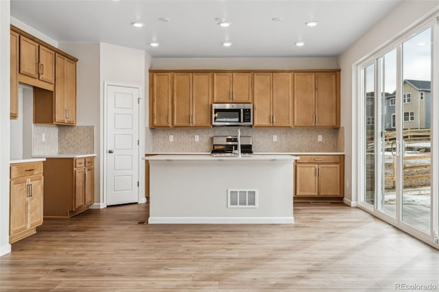 kitchen featuring stainless steel appliances, a center island with sink, light wood-type flooring, and decorative backsplash