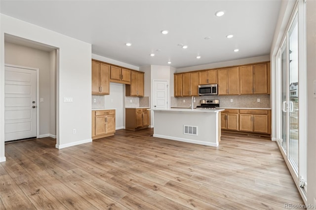 kitchen with sink, backsplash, stainless steel appliances, light hardwood / wood-style floors, and an island with sink