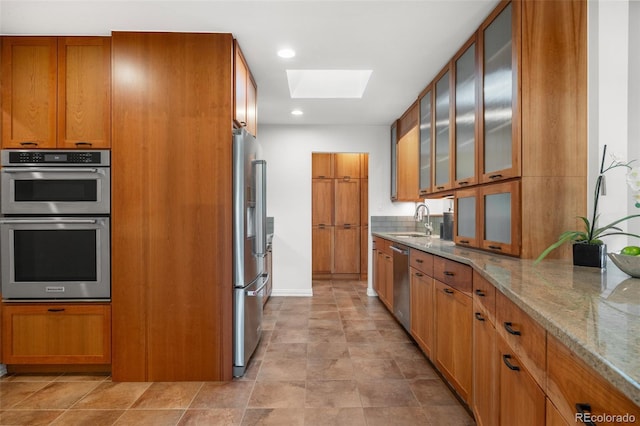 kitchen with light stone countertops, appliances with stainless steel finishes, a sink, brown cabinetry, and a skylight