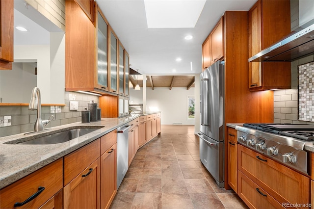 kitchen with brown cabinetry, light stone countertops, range hood, a sink, and appliances with stainless steel finishes