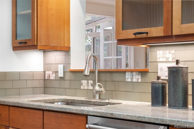 kitchen with tasteful backsplash, stainless steel dishwasher, brown cabinets, a sink, and light stone counters