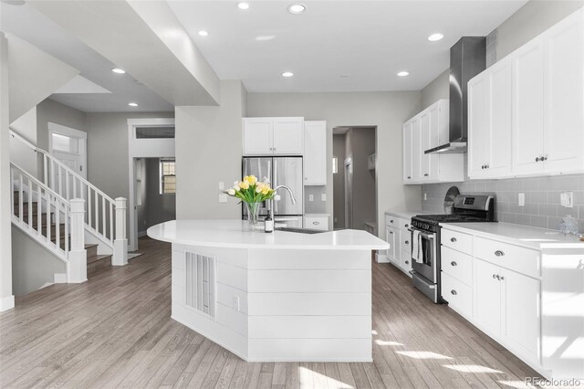 kitchen featuring stainless steel appliances, wall chimney range hood, light hardwood / wood-style floors, a center island with sink, and white cabinets