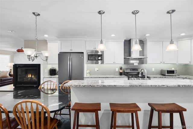 kitchen featuring appliances with stainless steel finishes, wall chimney exhaust hood, decorative light fixtures, and light stone counters