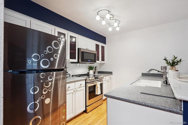 kitchen featuring appliances with stainless steel finishes, a sink, glass insert cabinets, white cabinets, and light wood-type flooring
