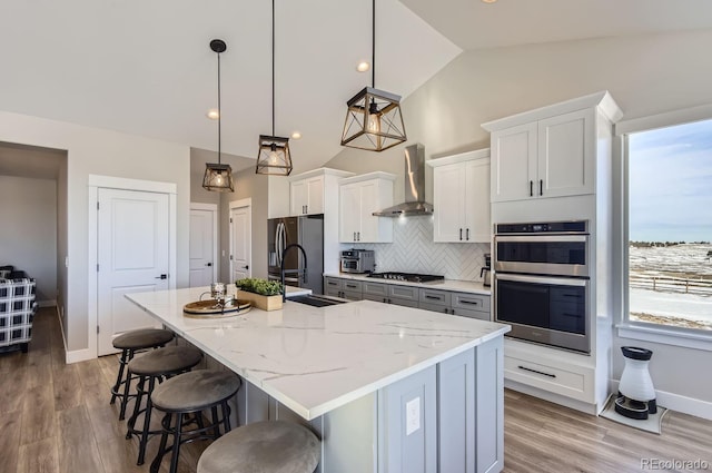 kitchen with pendant lighting, white cabinets, wall chimney range hood, stainless steel appliances, and an island with sink