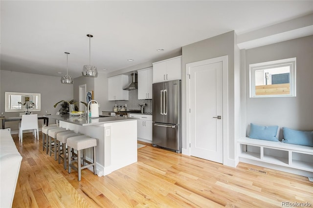 kitchen with high quality fridge, decorative light fixtures, white cabinets, a center island with sink, and wall chimney exhaust hood