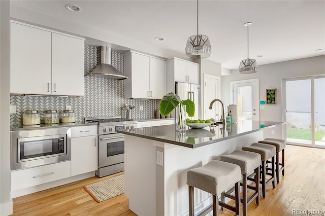 kitchen with white cabinetry, wall chimney range hood, hanging light fixtures, stainless steel appliances, and a center island with sink