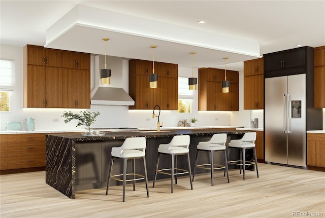 kitchen with light hardwood / wood-style flooring, wall chimney range hood, a center island with sink, high end refrigerator, and a breakfast bar area