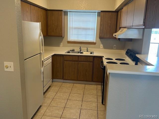 kitchen with brown cabinetry, a sink, light tile patterned floors, under cabinet range hood, and white appliances