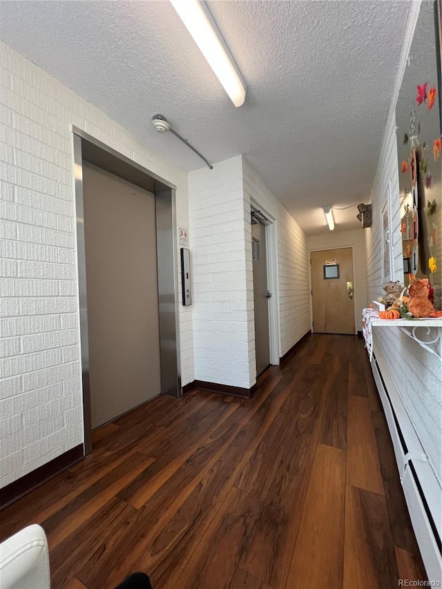 hallway with a textured ceiling, elevator, baseboard heating, dark hardwood / wood-style flooring, and brick wall