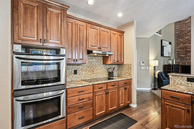 kitchen with stainless steel double oven, black electric cooktop, under cabinet range hood, dark wood-style flooring, and decorative backsplash