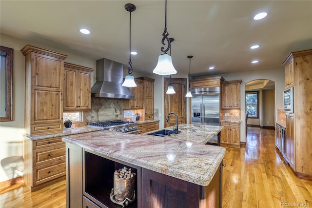 kitchen featuring backsplash, a spacious island, wall chimney range hood, built in appliances, and decorative light fixtures