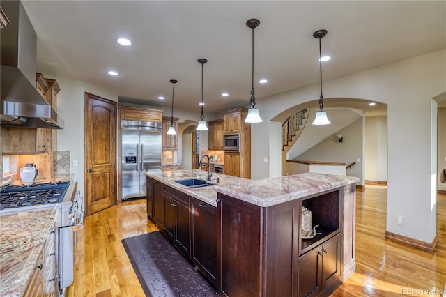 kitchen with a center island with sink, wall chimney range hood, sink, hanging light fixtures, and built in appliances