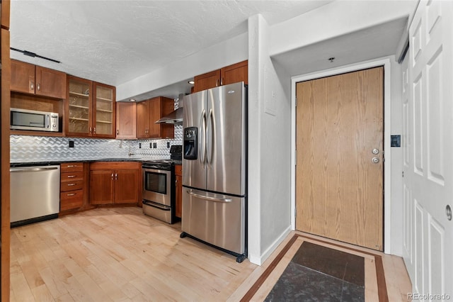 kitchen with wall chimney range hood, brown cabinets, tasteful backsplash, light wood-style flooring, and stainless steel appliances