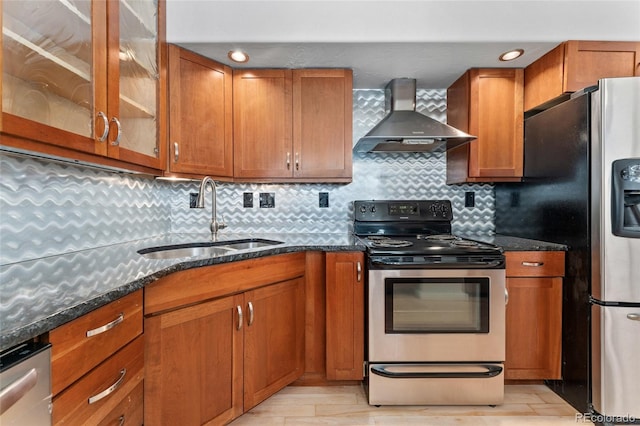 kitchen with brown cabinets, a sink, wall chimney exhaust hood, dark stone counters, and stainless steel appliances