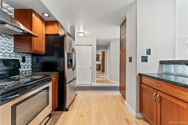 kitchen featuring wall chimney range hood, brown cabinets, stainless steel range with electric stovetop, light wood-style floors, and dark stone counters