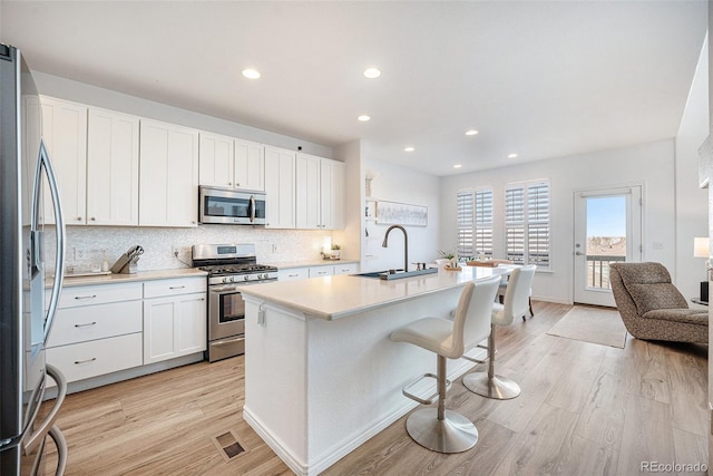 kitchen with white cabinets, stainless steel appliances, sink, a kitchen island with sink, and light wood-type flooring