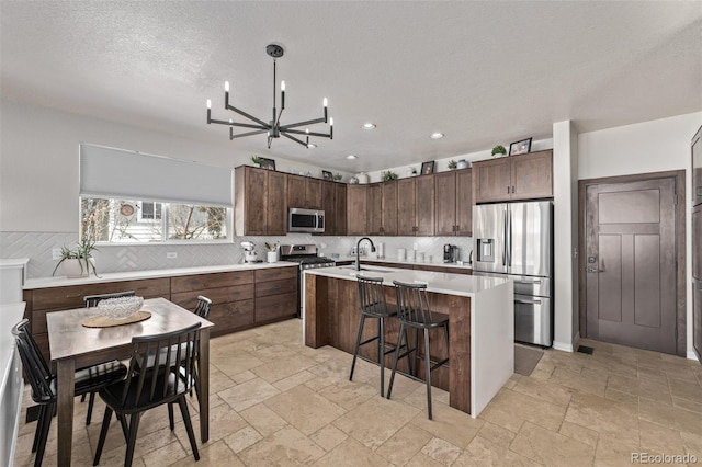 kitchen featuring a center island with sink, stone tile flooring, appliances with stainless steel finishes, light countertops, and decorative backsplash