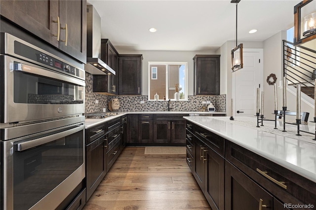 kitchen featuring wall chimney range hood, decorative backsplash, light wood-type flooring, pendant lighting, and stainless steel appliances