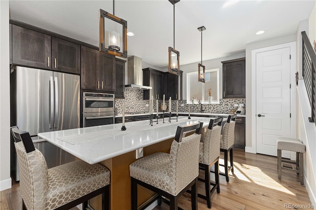 kitchen with a kitchen island with sink, decorative light fixtures, light hardwood / wood-style flooring, wall chimney range hood, and stainless steel appliances