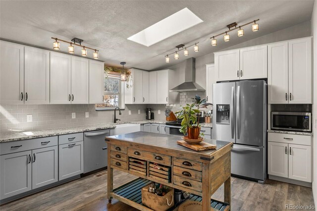 kitchen featuring wood finished floors, a sink, stainless steel appliances, lofted ceiling with skylight, and wall chimney range hood