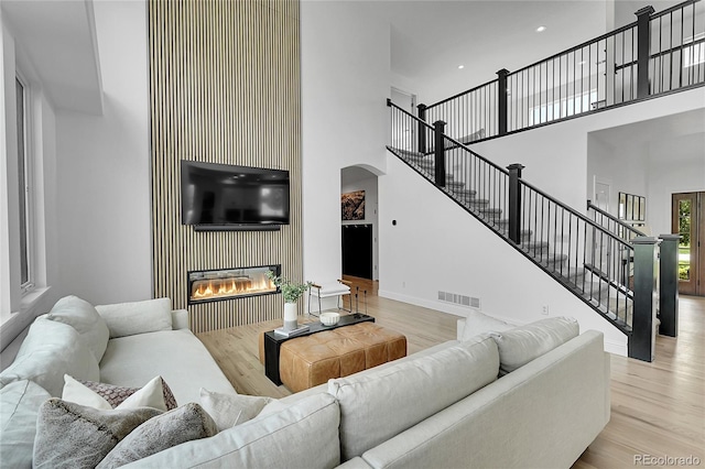 living area featuring a high ceiling, light wood-type flooring, arched walkways, a glass covered fireplace, and stairway