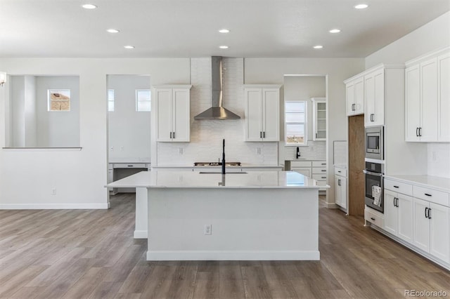 kitchen featuring white cabinetry, hardwood / wood-style flooring, stainless steel appliances, a kitchen island with sink, and wall chimney range hood