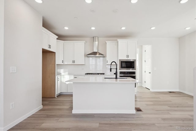 kitchen with white cabinetry, built in microwave, an island with sink, and wall chimney range hood