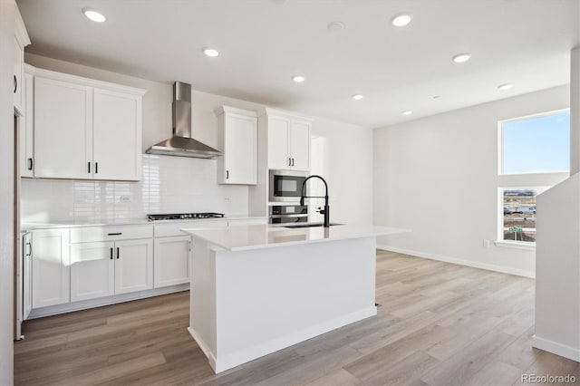 kitchen featuring white cabinetry, a kitchen island with sink, wall chimney range hood, and light wood-type flooring