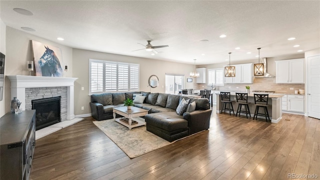 living room with a wealth of natural light, a stone fireplace, wood finished floors, and ceiling fan with notable chandelier