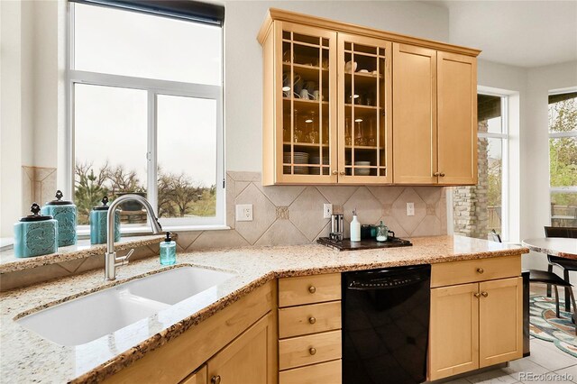 kitchen featuring black dishwasher, a sink, light brown cabinets, light stone counters, and glass insert cabinets