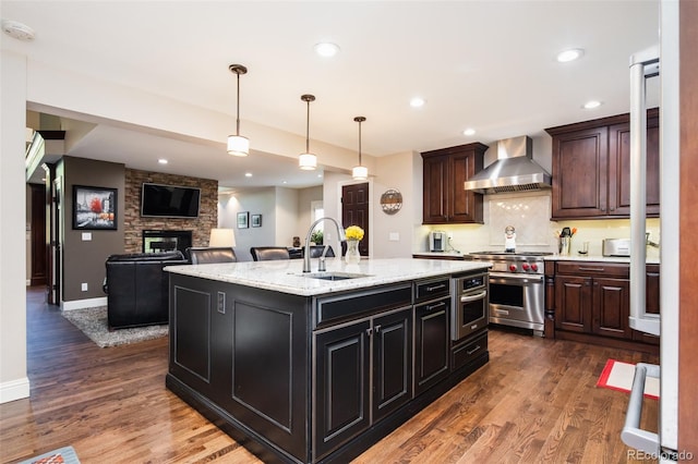 kitchen featuring dark wood-style floors, appliances with stainless steel finishes, a kitchen island with sink, wall chimney range hood, and a sink