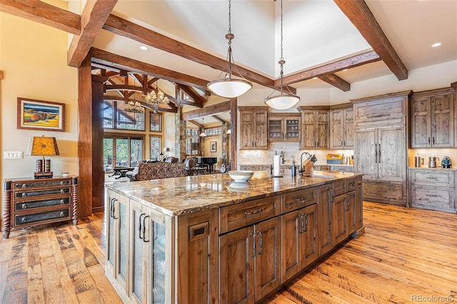 kitchen with light wood-type flooring, decorative light fixtures, a center island with sink, stone countertops, and beamed ceiling