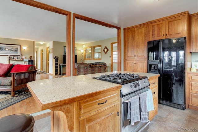 kitchen with black refrigerator with ice dispenser, brown cabinetry, a kitchen island, stainless steel range with gas stovetop, and light countertops