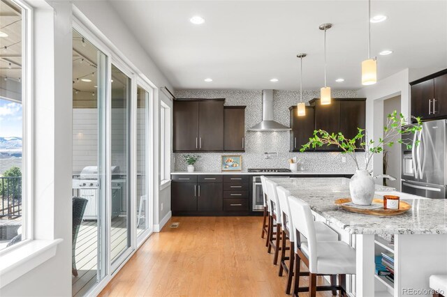 kitchen with dark brown cabinetry, wall chimney range hood, appliances with stainless steel finishes, a kitchen bar, and decorative light fixtures