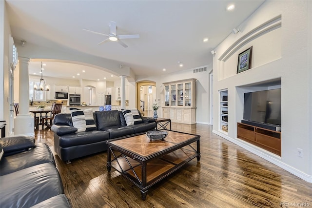 living room featuring decorative columns, built in shelves, ceiling fan with notable chandelier, and dark hardwood / wood-style flooring