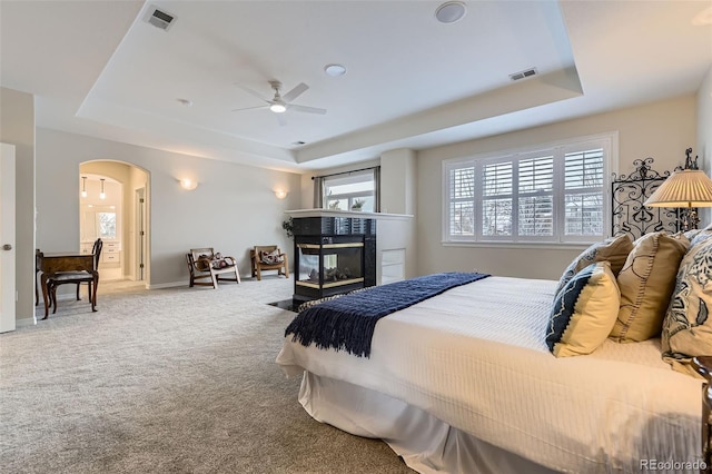 carpeted bedroom with ceiling fan, a tiled fireplace, and a tray ceiling